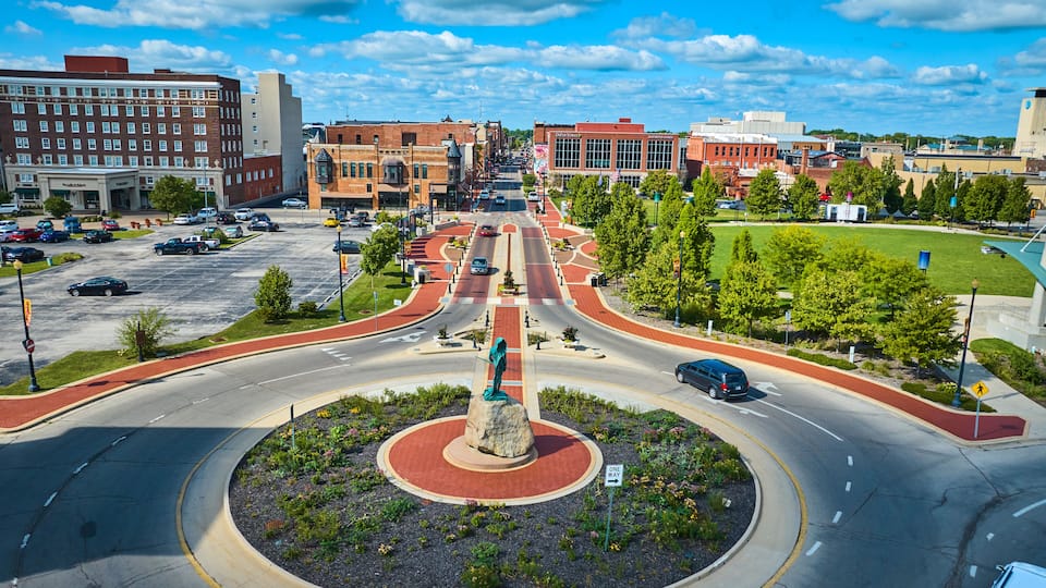A city intersection with a central monument, multiple lanes, and surrounding buildings.
