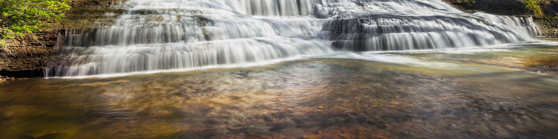 Uma cascata em vários níveis numa área arborizada, com águas cristalinas e vegetação exuberante ao redor.