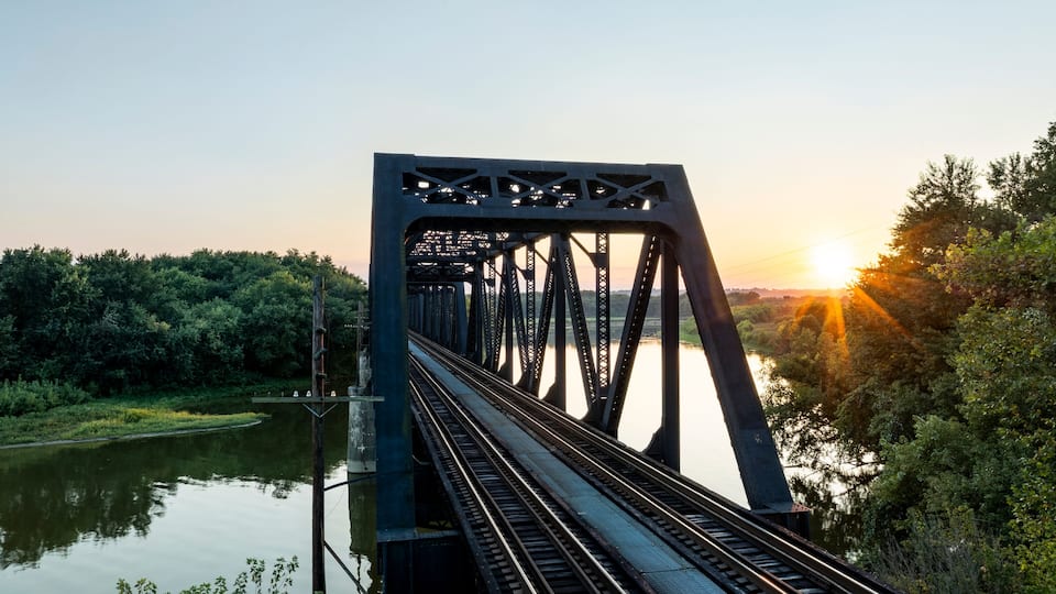 A steel bridge with train tracks over a calm river, surrounded by dense greenery and a sunset in the background.