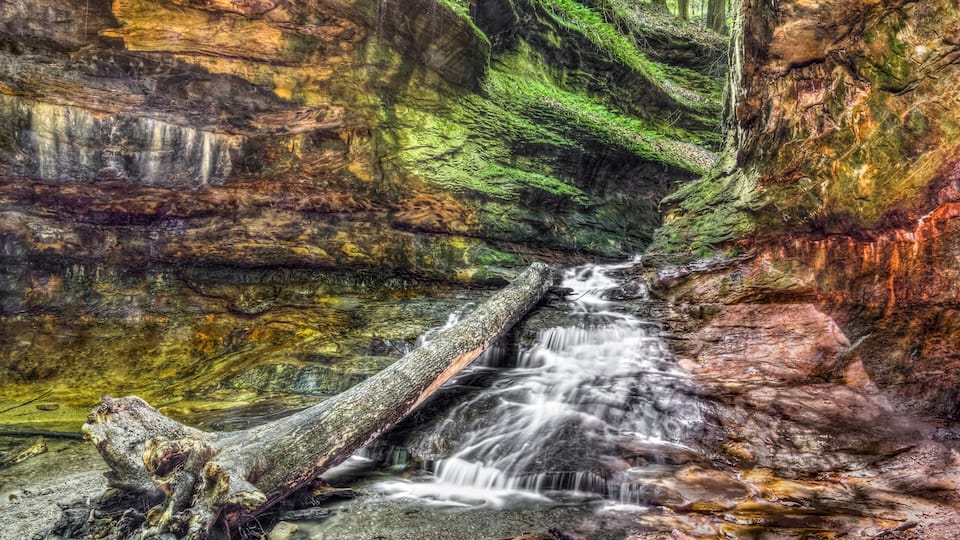 Water cascades over colorful sandstone from a narrow passage with green rocky walls In Indiana's Turkey Run State Park.