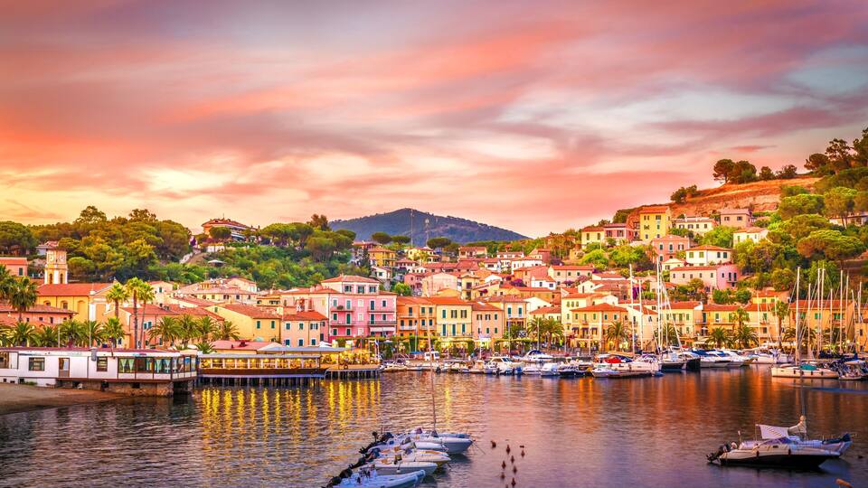 A coastal town with colorful buildings, a marina with boats, and a hill in the background.