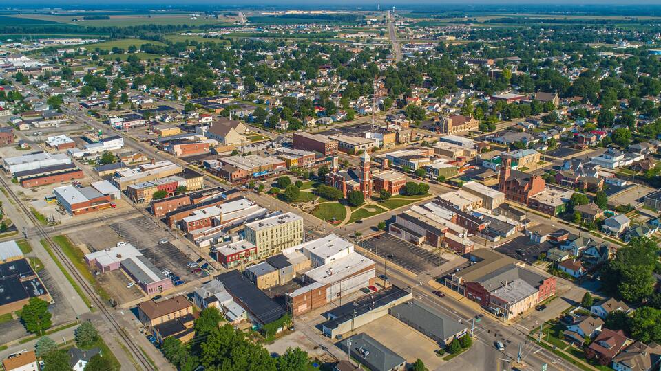 An aerial view of a small town with a mix of residential and commercial buildings, a prominent church steeple, and a bridge in the distance.