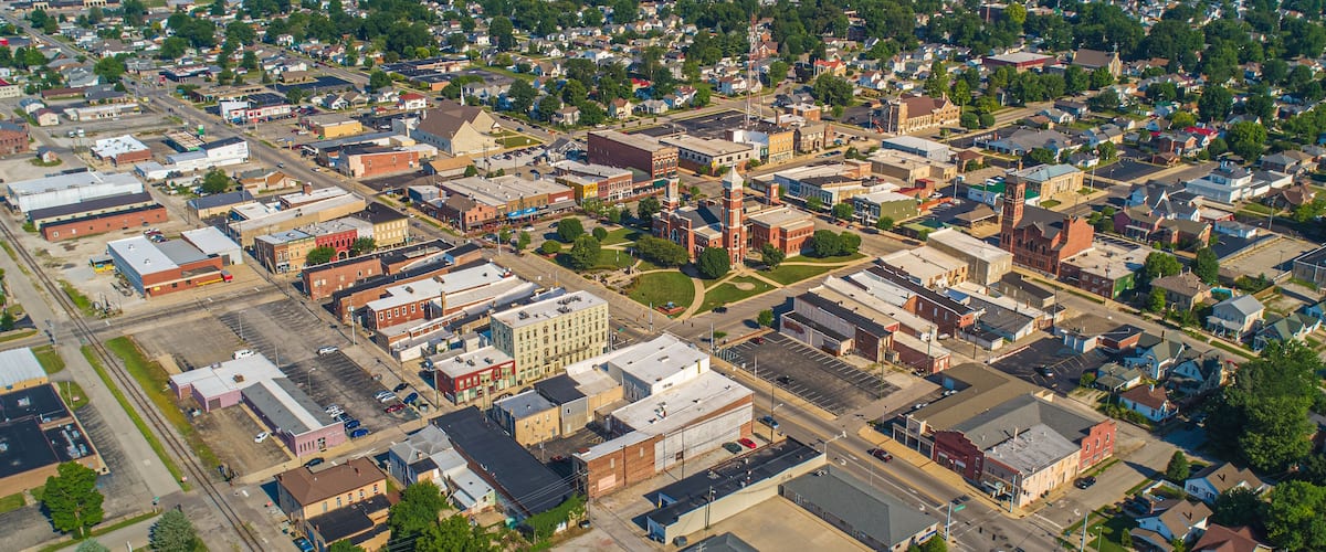 An aerial view of a small town with a mix of residential and commercial buildings, a prominent church steeple, and a bridge in the distance.