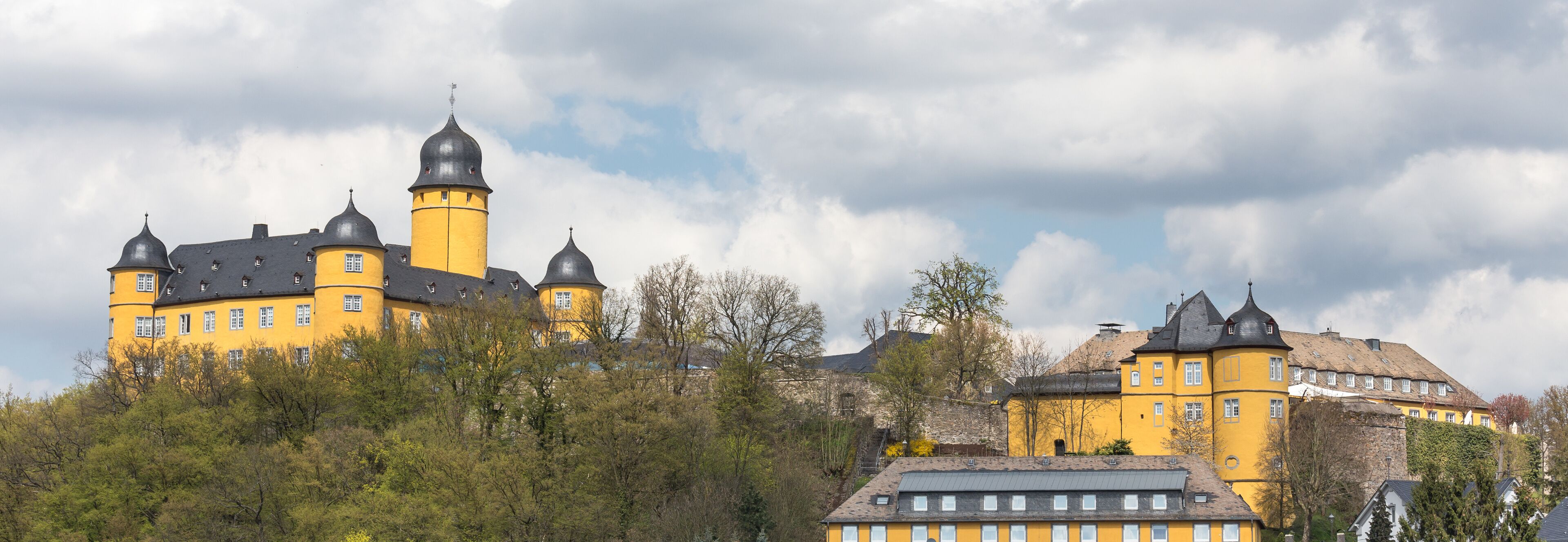 A yellow castle with multiple towers and a large central tower, surrounded by trees and other buildings.