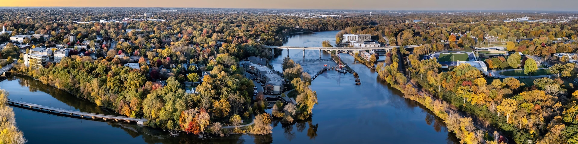 An aerial view of a river with a bridge, surrounded by autumn foliage and urban buildings.