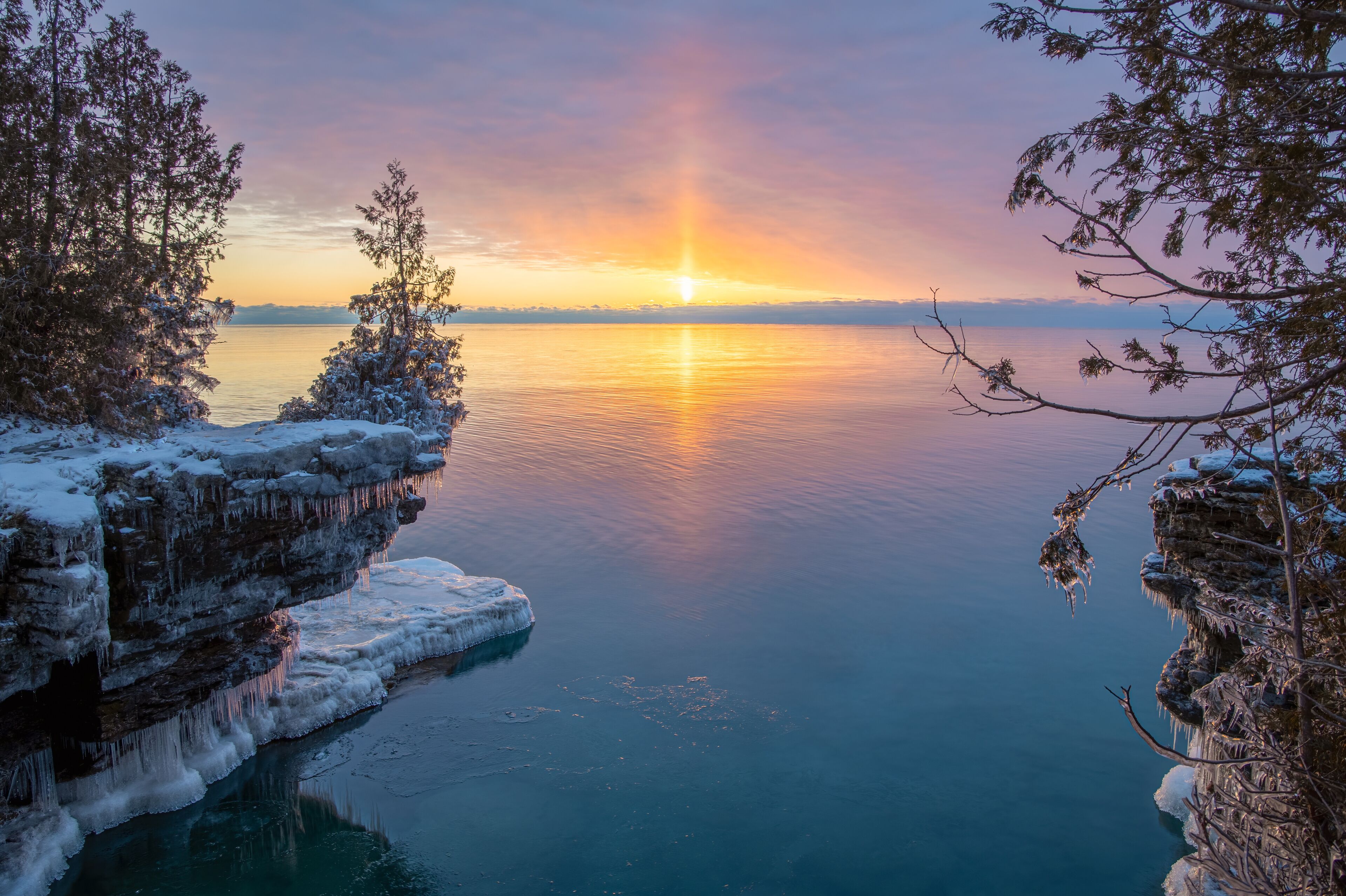 Un lago helado con estalactitas, un atardecer y árboles cubiertos de nieve.