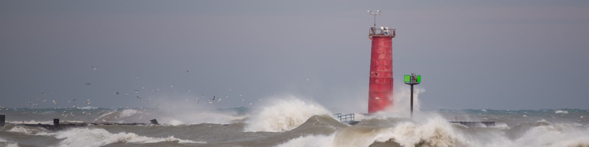 A red lighthouse amidst rough seas with waves crashing against a pier.