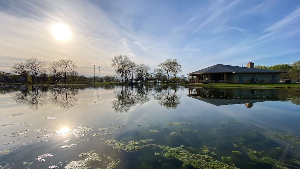 A serene lake with a clear reflection of trees and a building, under a bright sun.