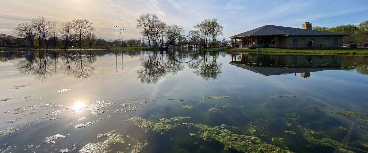 A serene lake with a clear reflection of trees and a building, under a bright sun.