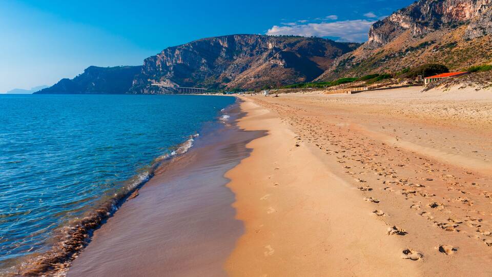 A beach with clear blue water, a sandy shore, and rocky cliffs in the background.