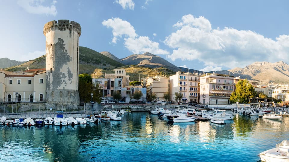 A coastal town with a historic tower, a marina with boats, and mountains in the background.