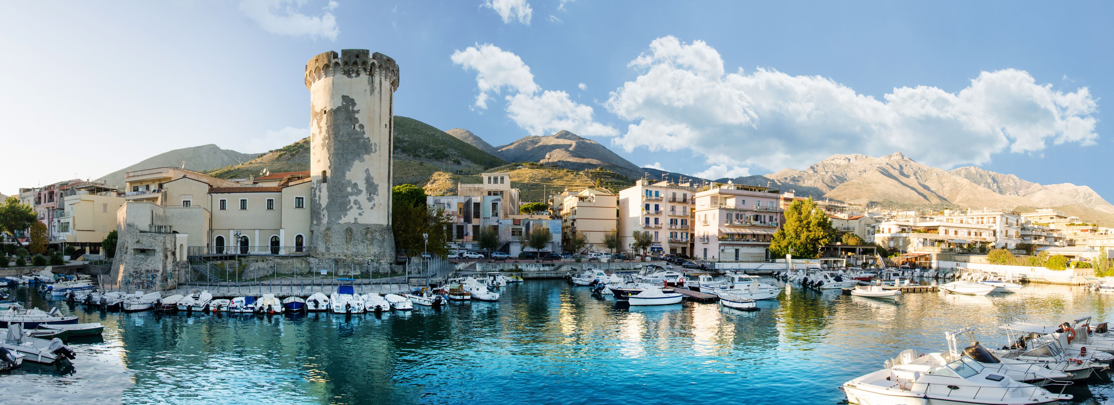 A coastal town with a historic tower, a marina with boats, and mountains in the background.