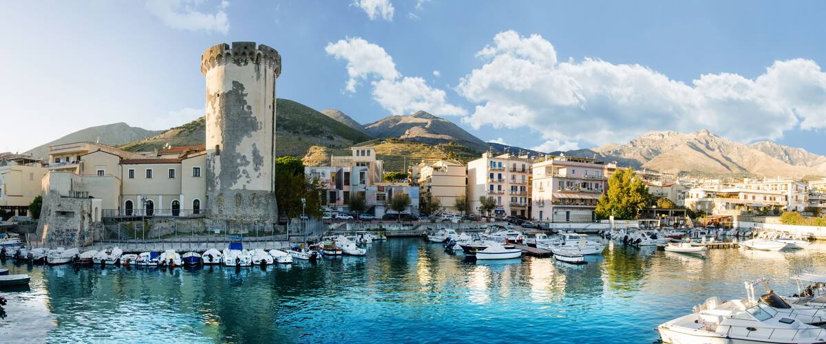 A coastal town with a historic tower, a marina with boats, and mountains in the background.