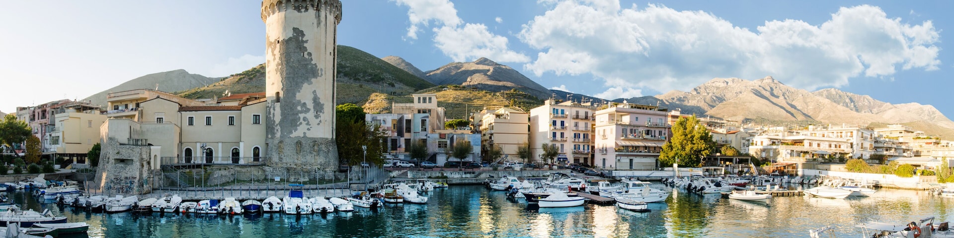 A coastal town with a historic tower, a marina with boats, and mountains in the background.