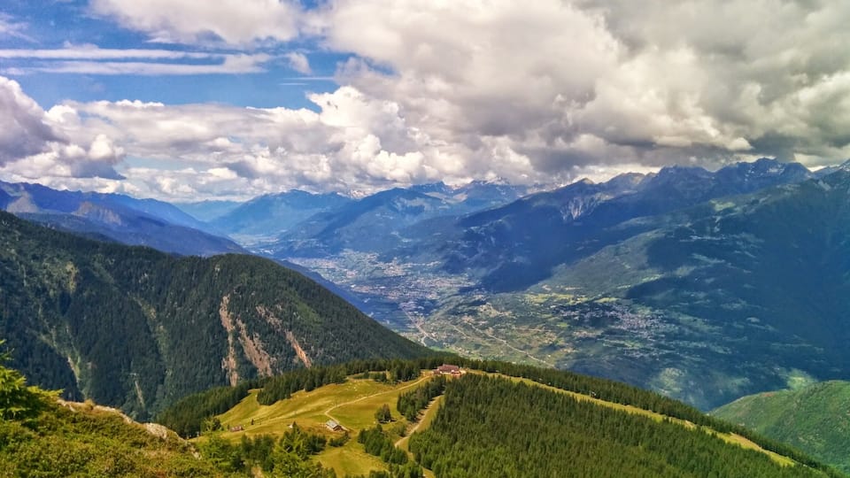 A mountainous landscape with a valley, forested hills, and distant peaks under a partly cloudy sky.