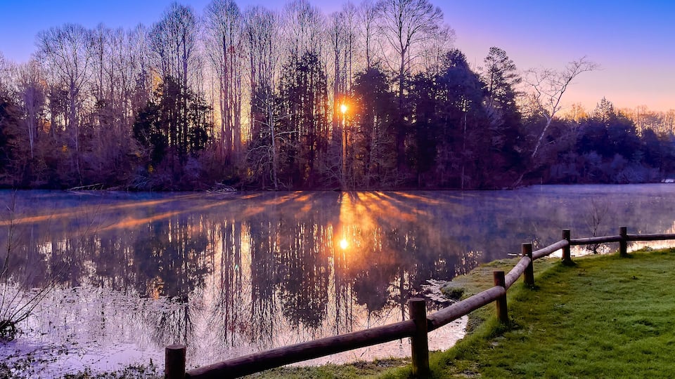 A serene lakeside scene at dusk with a wooden fence, trees, and a street lamp reflecting on the water.
