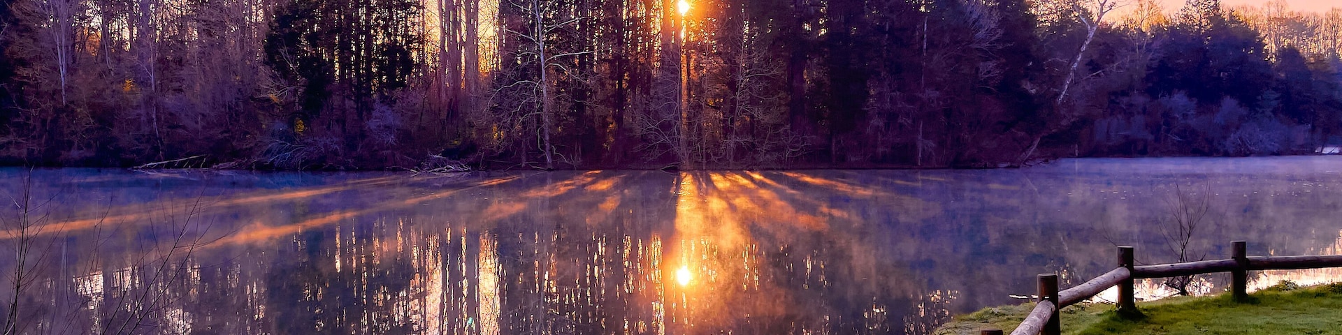 Escena serena a orillas de un lago al anochecer, con una valla de madera, árboles y un farol que se refleja en el agua.
