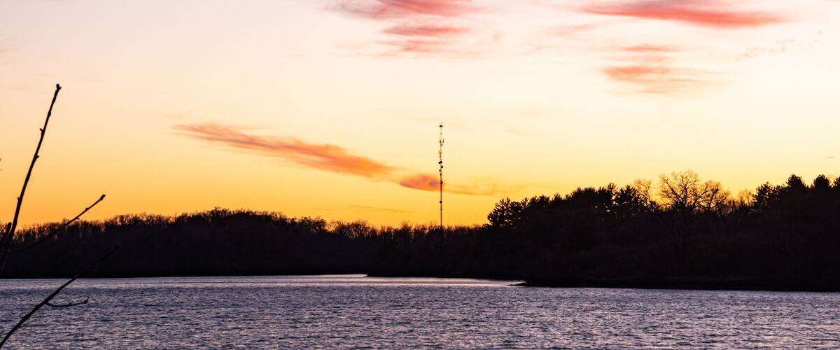 A sunset over a body of water with a silhouette of trees and a communication tower.