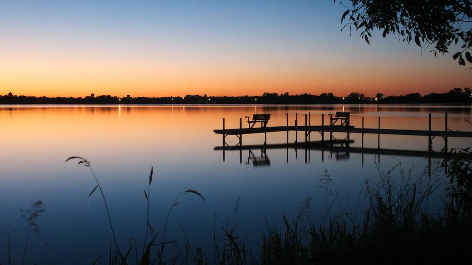 A calm lake at sunset with a wooden dock and benches.