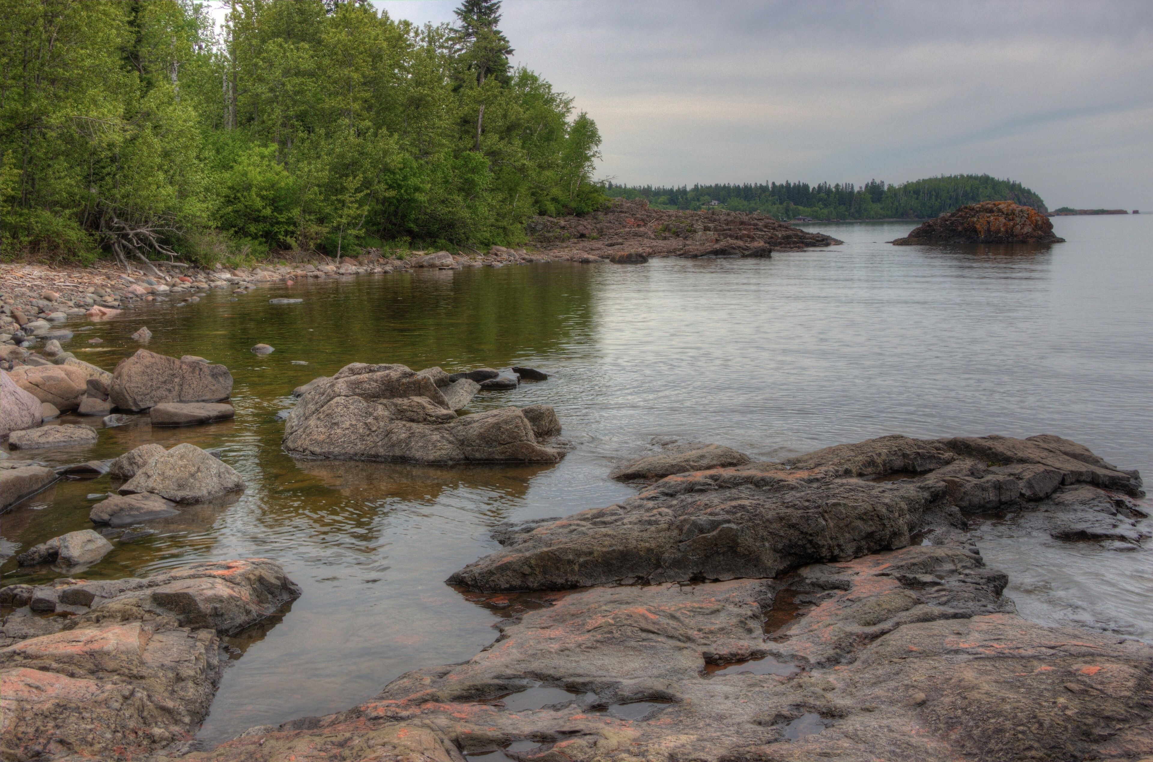 A calm lake with large rocks in the foreground and dense forest on the left.