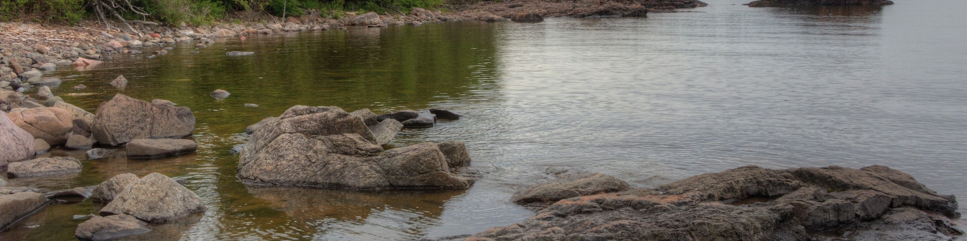 A calm lake with large rocks in the foreground and dense forest on the left.