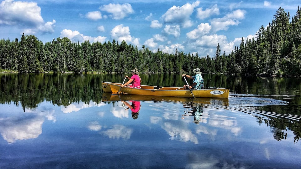 Two people canoeing on a calm lake surrounded by dense forest.
