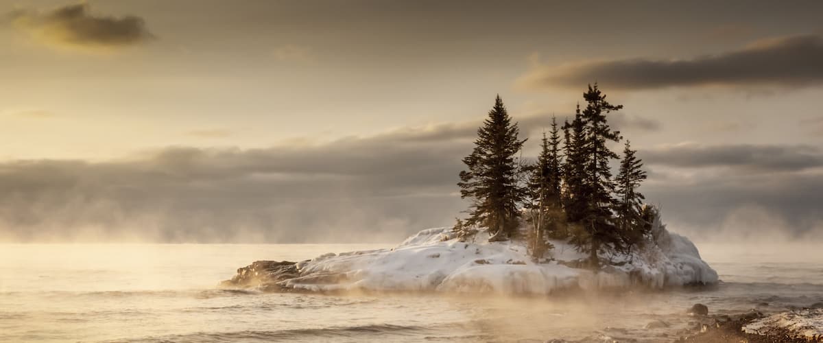 A snow-covered island with two tall evergreen trees and a misty ocean background.
