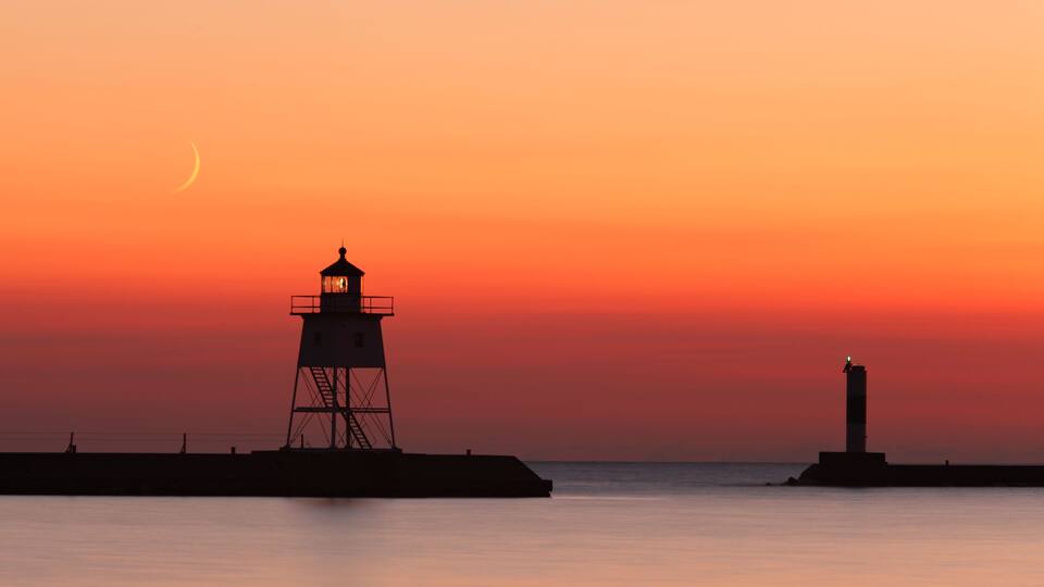 A lighthouse silhouette against a sunset sky, calm water, and a crescent moon.