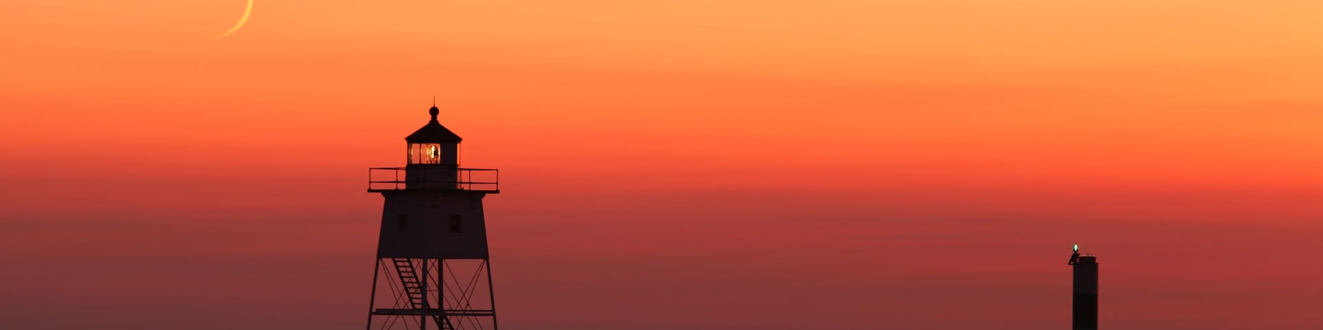 Silueta de un faro contra un cielo de atardecer, agua tranquila y una luna creciente.
