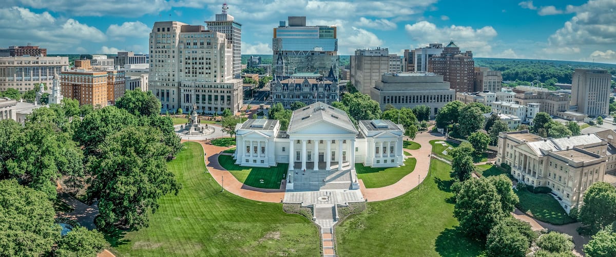 A cityscape with a prominent white building with columns, surrounded by lush green lawns and trees, under a partly cloudy sky.