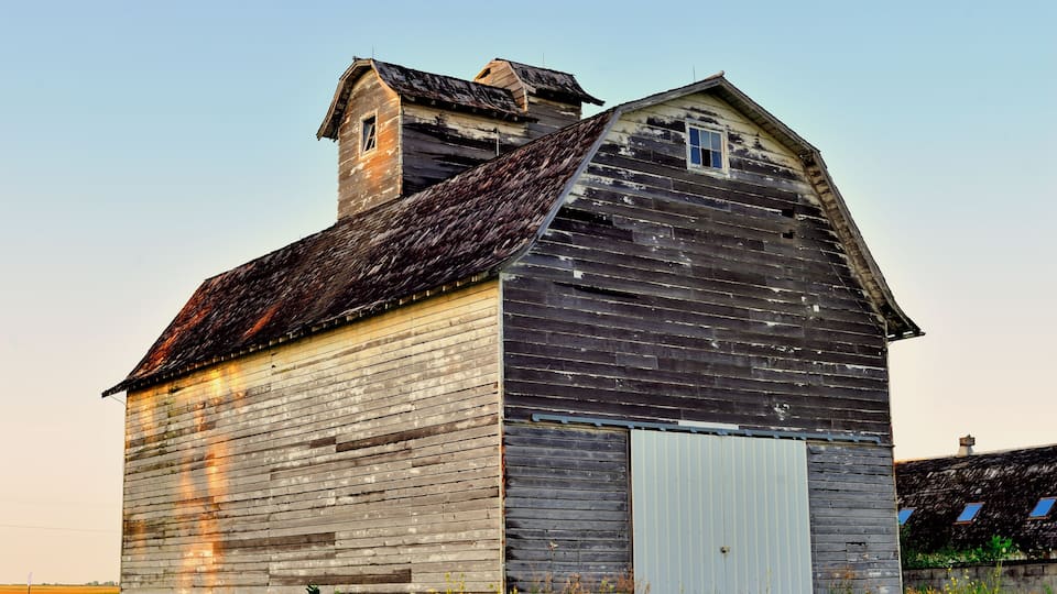 An old wooden barn with a sloped roof and a small window, surrounded by a grassy field.