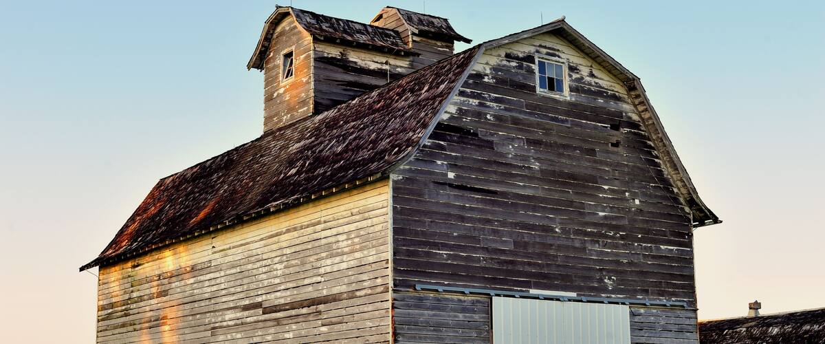 An old wooden barn with a sloped roof and a small window, surrounded by a grassy field.
