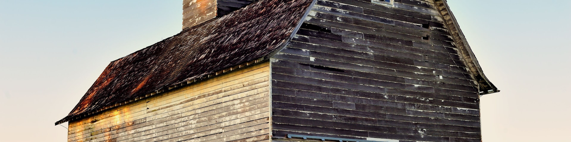 An old wooden barn with a sloped roof and a small window, surrounded by a grassy field.