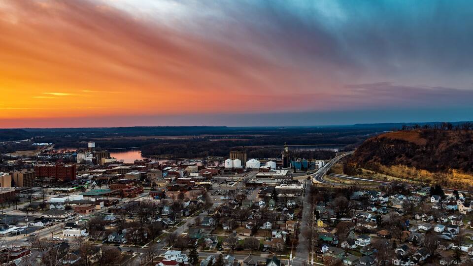 A panoramic view of a town at sunset with a river, industrial buildings, and a bridge.