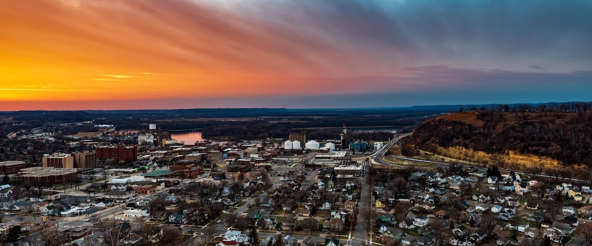 A panoramic view of a town at sunset with a river, industrial buildings, and a bridge.