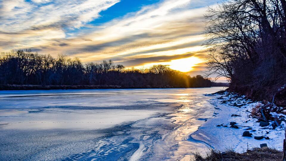 A frozen river with a sunset, bare trees, and snow on the banks.