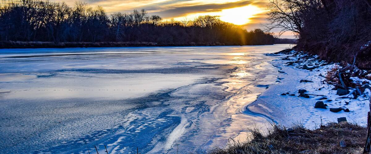 A frozen river with a sunset, bare trees, and snow on the banks.