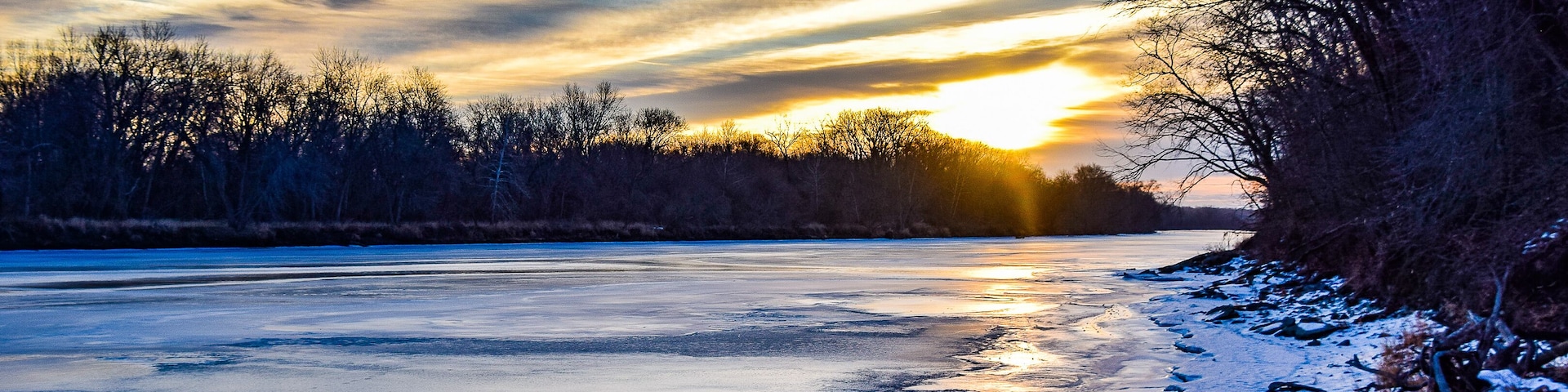 A frozen river with a sunset, bare trees, and snow on the banks.