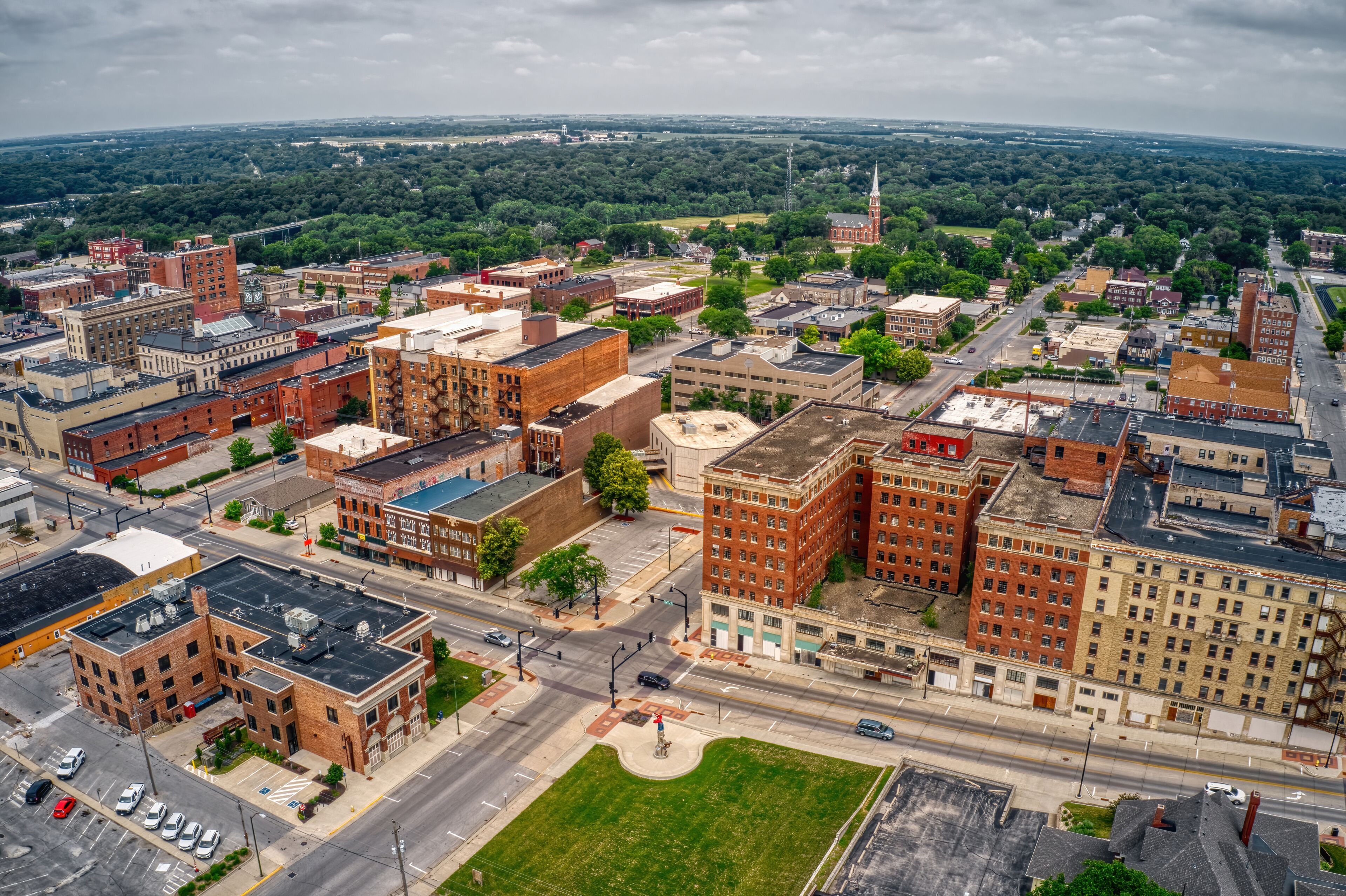 An aerial view of a city with a prominent red-brick building, a green park, and a distant church steeple.