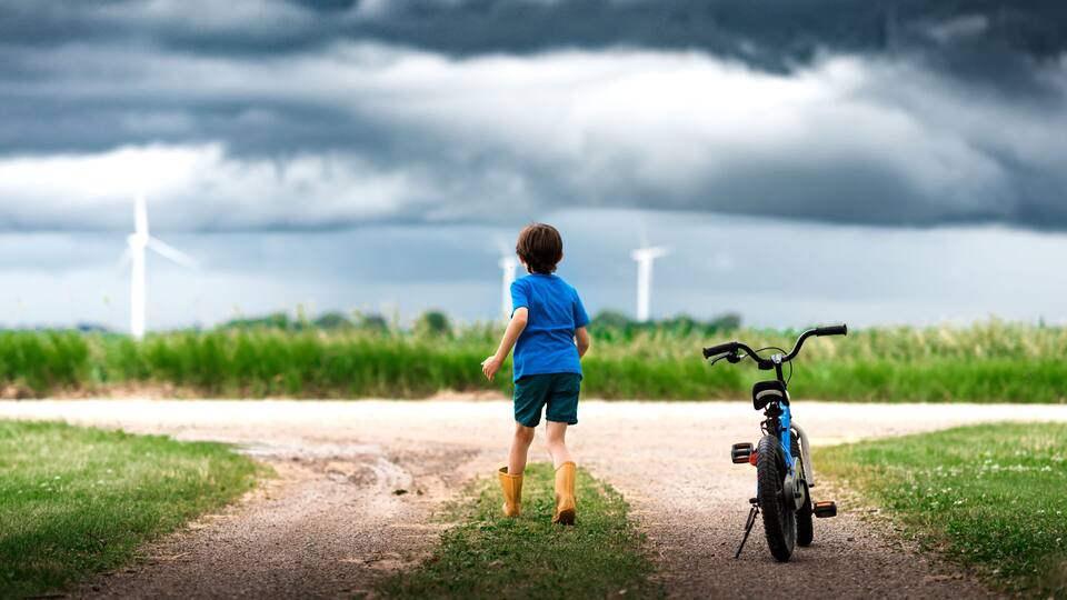 A child walking on a dirt path with a bicycle, wind turbines in the background, and a cloudy sky.