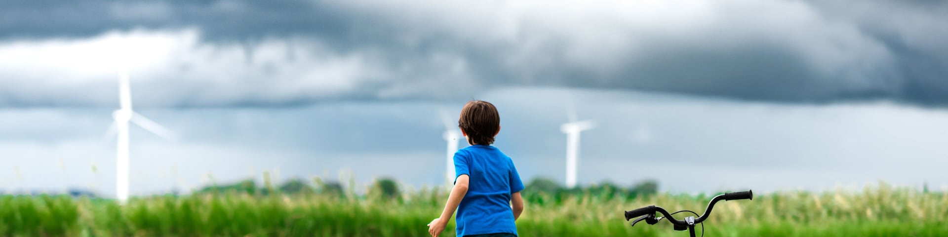 A child walking on a dirt path with a bicycle, wind turbines in the background, and a cloudy sky.