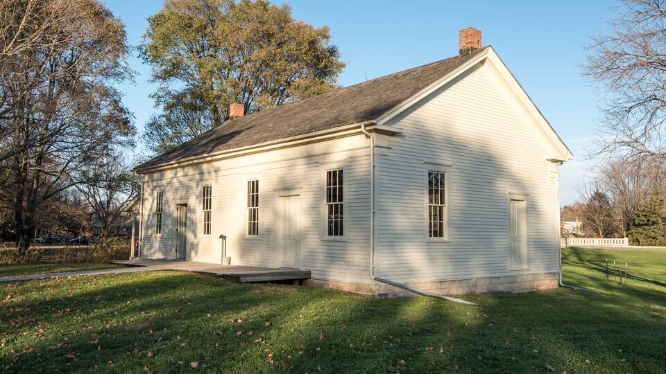 A white wooden house with a gabled roof and a chimney, surrounded by a grassy lawn and trees.