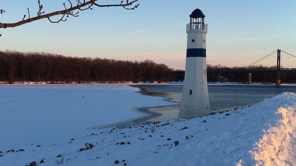 A lighthouse on a snow-covered shore with a bridge in the background.