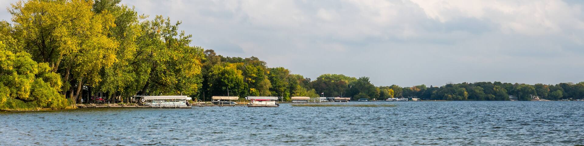 A calm lake with boats, trees, and a cloudy sky.