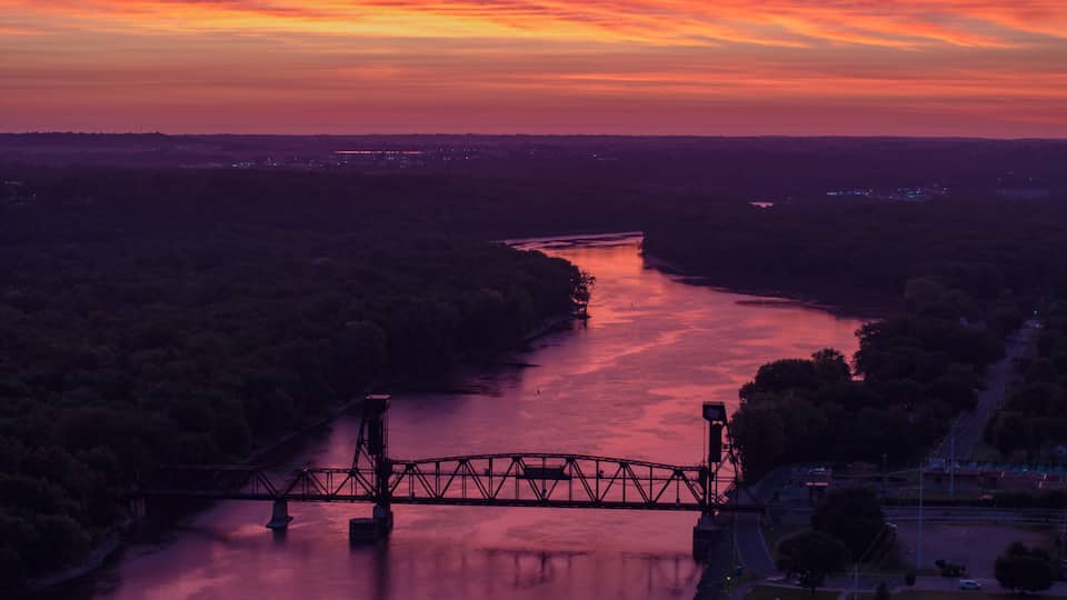 A sunset over a river with a bridge, surrounded by dense forest.
