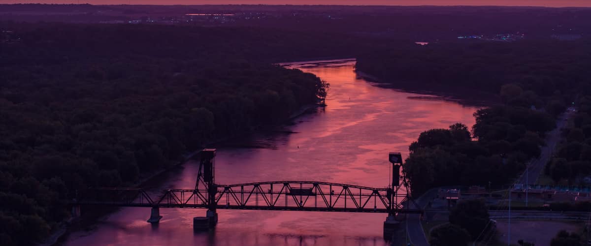 A sunset over a river with a bridge, surrounded by dense forest.