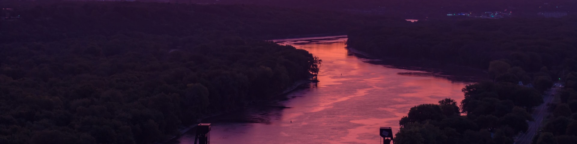 A sunset over a river with a bridge, surrounded by dense forest.