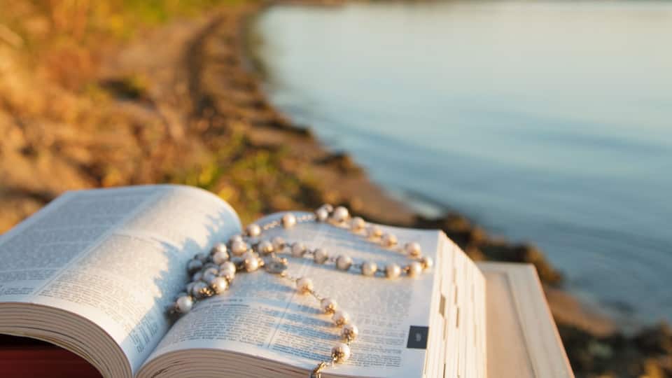 An open book with rosary beads on a wooden table by a calm lake.