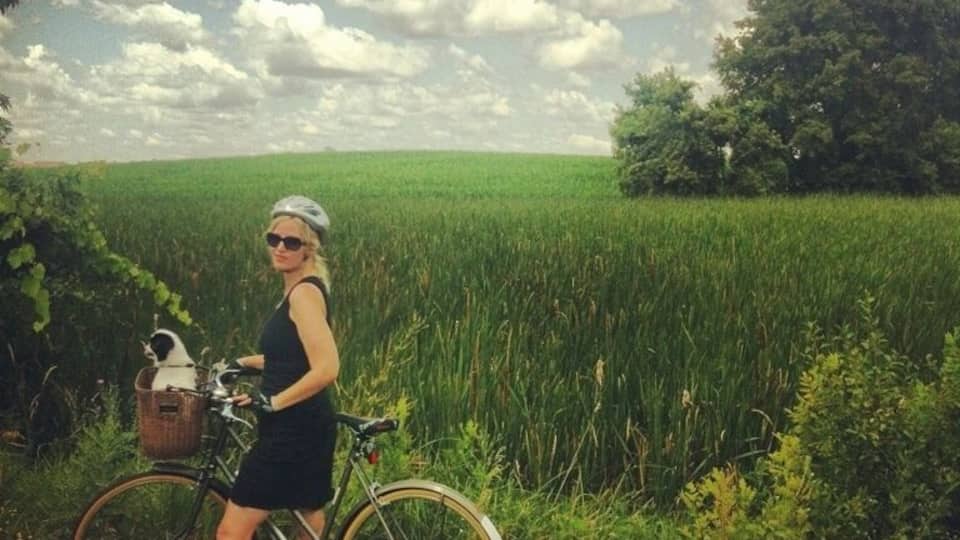 A woman standing next to a bicycle in a field with a basket containing a cat.