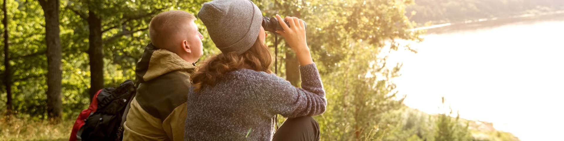 A person taking a photograph with a camera, wearing a hat and a sweater, standing near a body of water surrounded by trees.